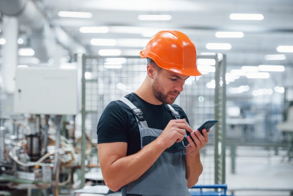 smartphone in hands. industrial worker indoors in factory. young technician with orange hard hat smartphone in hands. industrial worker indoors in factory. young technician with orange hard hat